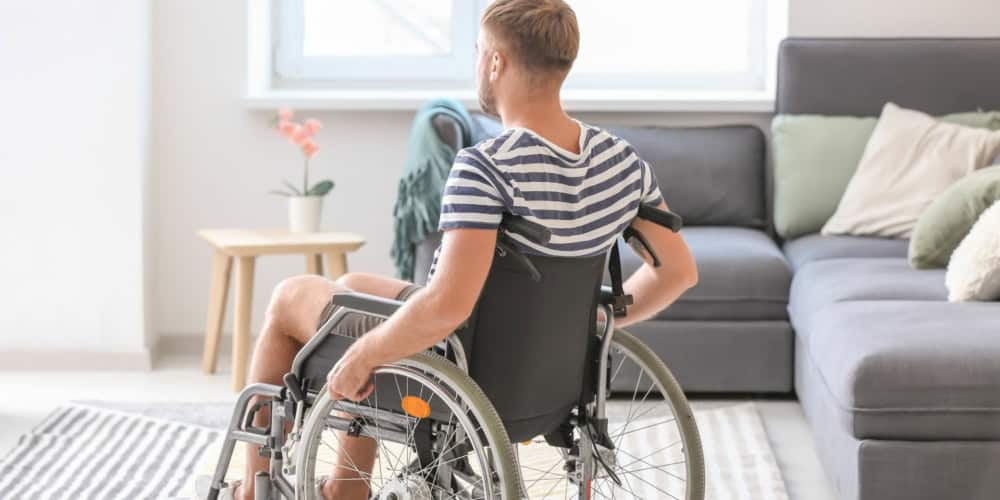 man in a wheelchair in a striped shirt, moving past his grey lounge in the living room. Facing away from the camera.